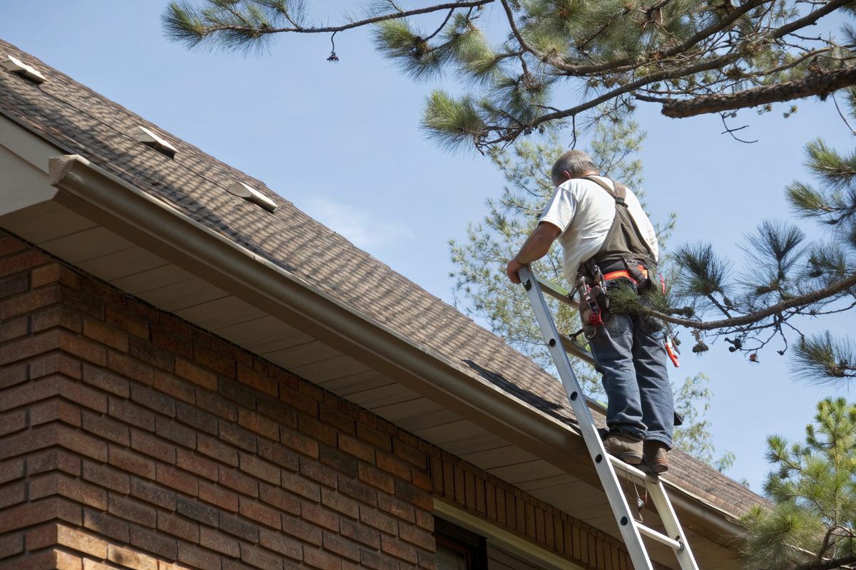 A roofer in a harness on a ladder inspecting the shingles and gutter of a brick house
